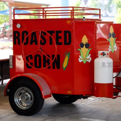 Bright red trailer with "ROASTED CORN" text, corn graphics, and propane tanks, parked under a shelter near a trash can and other vehicles.
