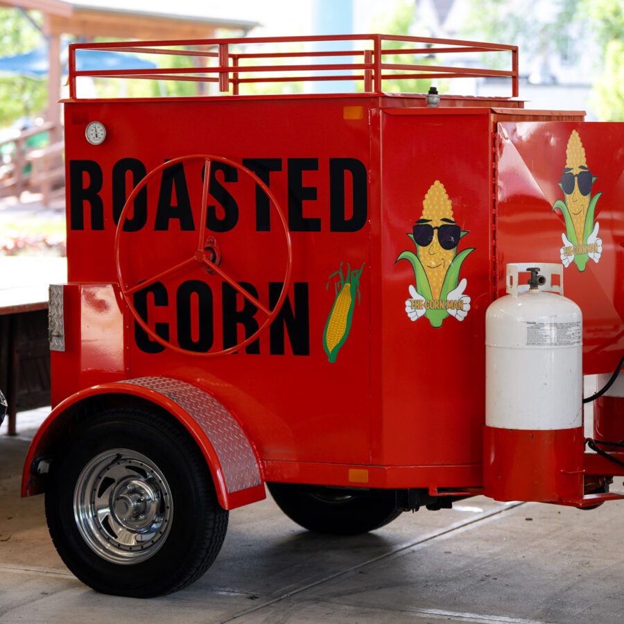 Bright red trailer with "ROASTED CORN" text, corn graphics, and propane tanks, parked under a shelter near a trash can and other vehicles.
