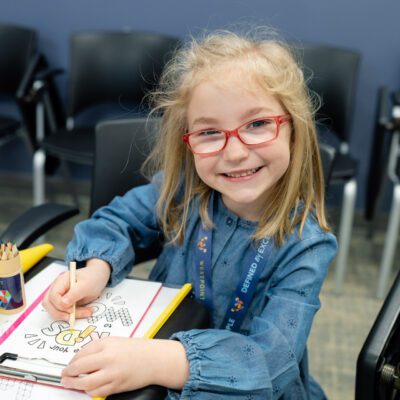 A young girl with glasses colors a drawing on paper at a desk, smiling at the camera. Colored pencils, a water bottle, and craft supplies are nearby.