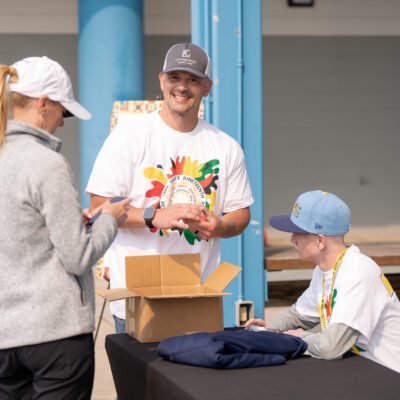 Four people interact at an outdoor event table; two wear white shirts and hats, one sits behind the table, and a woman in a gray jacket stands in front, using her phone.