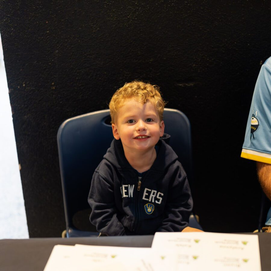 A young boy with curly hair sits on a chair at a table, smiling, with two adults partially visible beside him. Papers are spread out on the table in front.