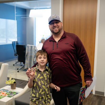 An adult and a child stand together smiling in a modern office lobby. The child holds a small potted plant and the adult holds colorful papers.