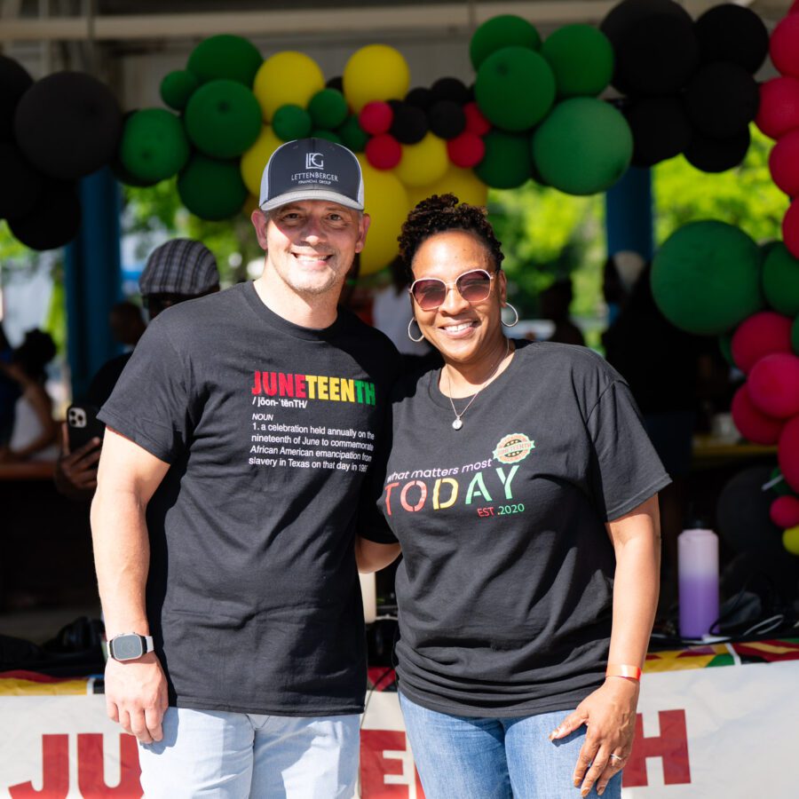 Two people stand smiling in front of a table decorated with Juneteenth-themed balloons and a banner at an outdoor event.