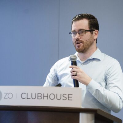 A man with glasses and a beard speaks into a microphone while standing at a podium labeled "CLUBHOUSE.