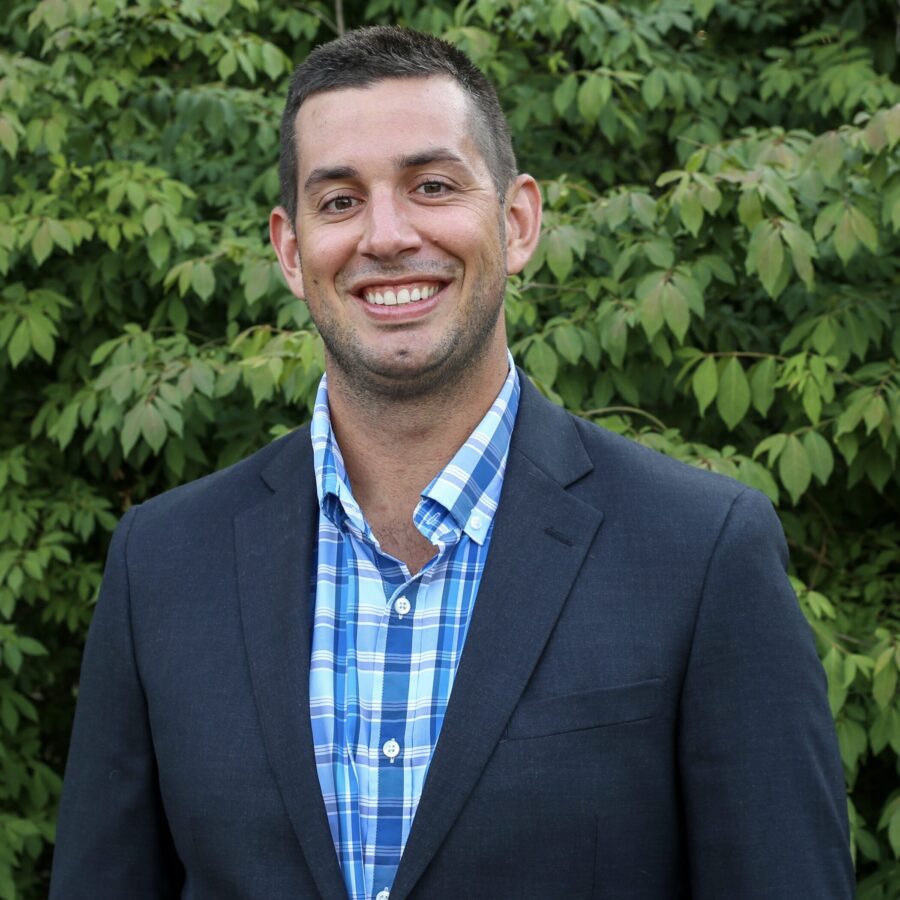 A man in a dark blazer and blue plaid shirt stands outdoors in front of green leafy bushes, smiling at the camera.