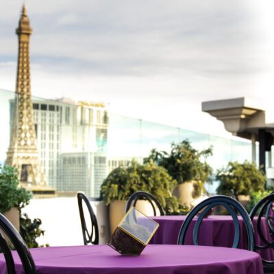 Outdoor restaurant tables covered with purple tablecloths overlook city buildings and a replica of the Eiffel Tower in the background.