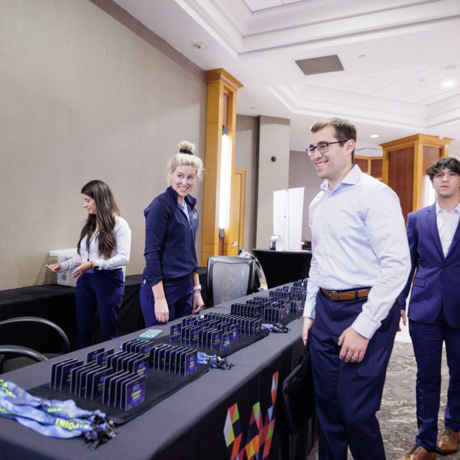 People in business attire stand around a registration table with name badges and lanyards at a conference or event venue.
