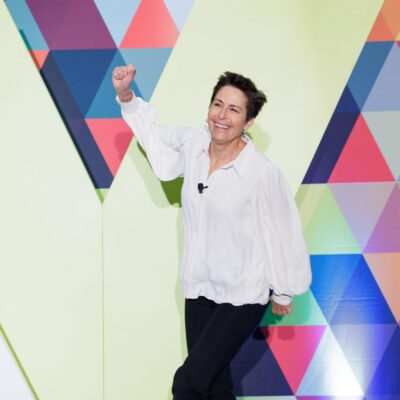 Person wearing a white blouse and black pants raises a fist and smiles while standing in front of a colorful geometric-patterned wall.