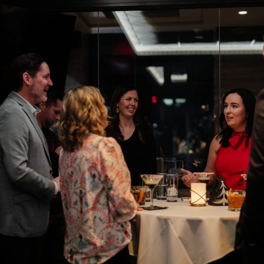 Five people stand around a cocktail table engaged in conversation in a dimly lit indoor setting. Drinks and a candle are on the table.