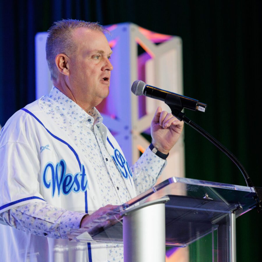 A man in a "West O" baseball jersey speaks at a podium with a microphone, gesturing with his right hand. Geometric décor and a projection screen are visible in the background.