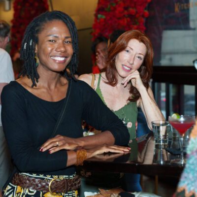 Three women are sitting and smiling at a bar table, with drinks in front of them; other people and red flowers are visible in the background.