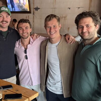 Four men stand together, smiling at a wooden bar table with drinks in a casual indoor setting.