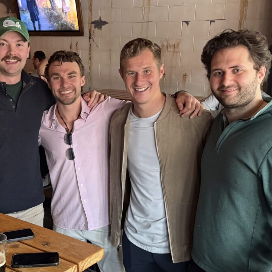 Four men stand together, smiling at a wooden bar table with drinks in a casual indoor setting.