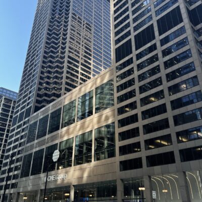 Tall modern office buildings with glass and steel facades line a city street; a black car is parked by the curb in front of One Gold building.