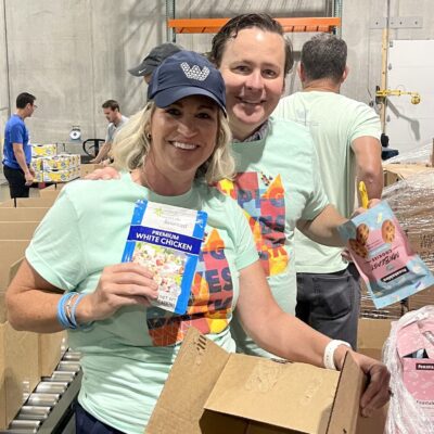 Two people in mint green shirts pack boxes with food items in a warehouse setting, smiling at the camera.