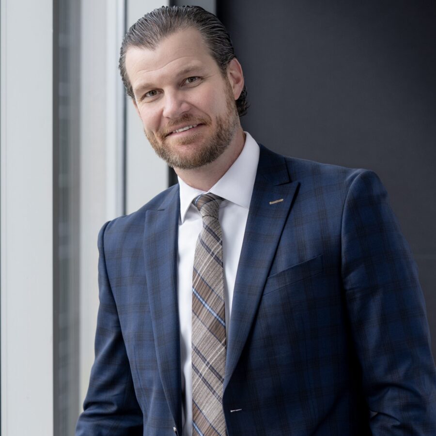 A man with short, slicked-back hair and a beard wears a blue plaid suit, white shirt, and striped tie, standing by a window with a neutral expression.
