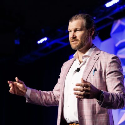 A man in a pink plaid jacket gestures while speaking on stage, wearing a microphone headset and standing under stage lights.