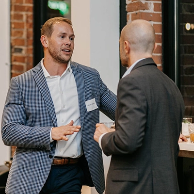Two men in business attire have a conversation at an indoor networking event, with drinks and exposed brick walls in the background.