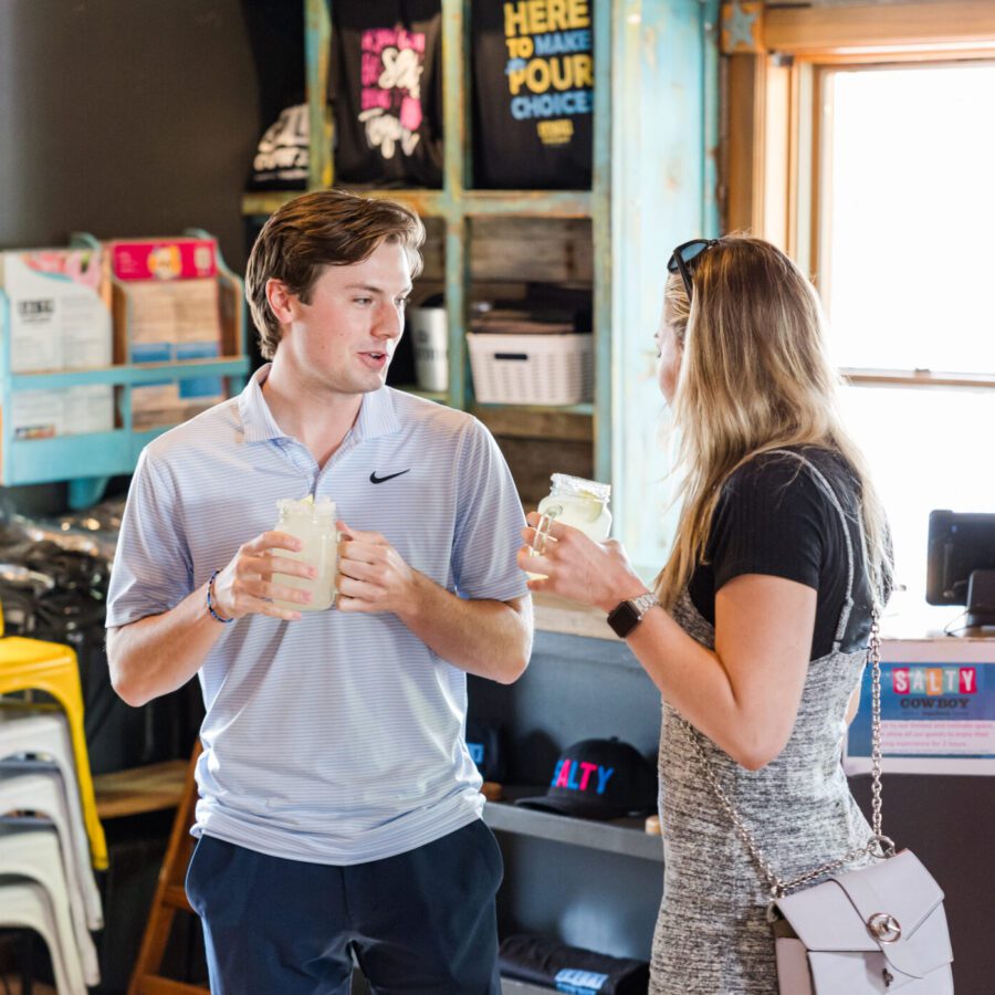 Two people stand indoors, each holding a drink in a mason jar, talking near stacked chairs and merchandise in a brightly lit shop.