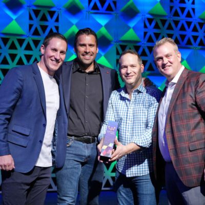 Four men stand together smiling as one holds a colorful award. They are dressed in business casual attire with a geometric pattern backdrop behind them.
