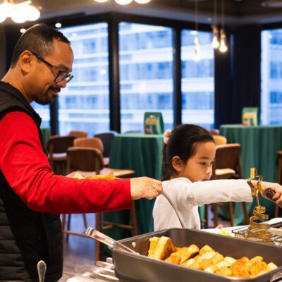 An adult and a child serve themselves food from a buffet in a brightly lit room with tables and large windows in the background.