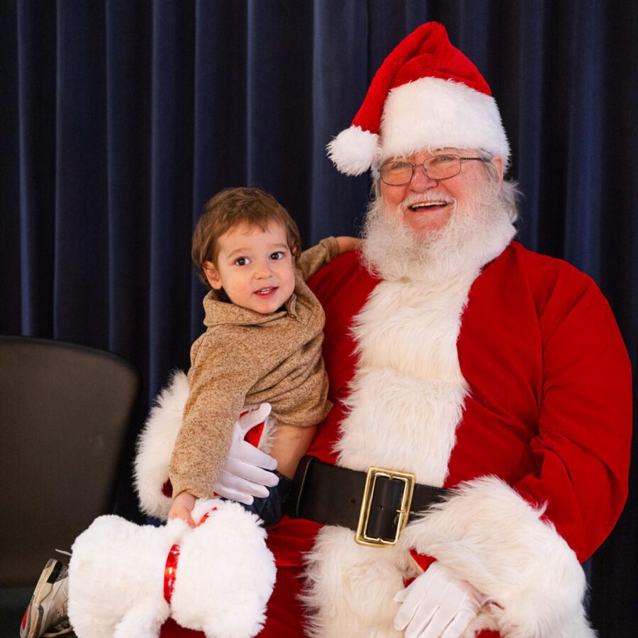 A child sits on Santa Claus’s lap, smiling at the camera, with a stuffed white dog toy in hand. Santa is dressed in a red suit and hat, sitting in front of a dark curtain.