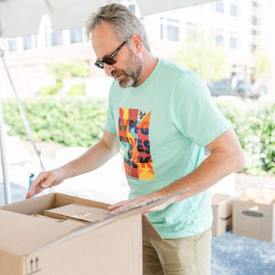 A man wearing sunglasses and a colorful t-shirt stands outdoors, opening a cardboard box on a table under a canopy.