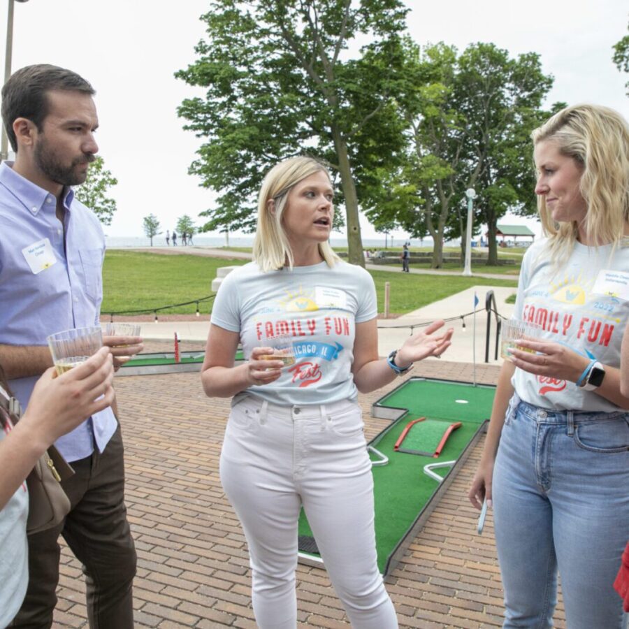 A group of five adults stands outside on a brick patio, talking and holding drinks near a mini-golf course. Trees and a lawn are visible in the background.
