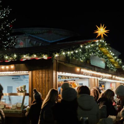 People gather at illuminated outdoor market stalls at night, with a decorated Christmas tree and star-shaped lights in the background.