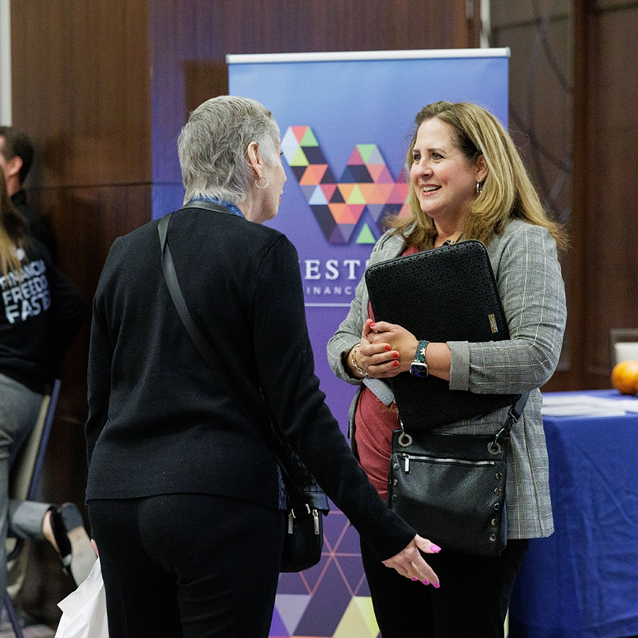Two women are talking in front of a booth with a geometric logo and the word "INVEST" on the backdrop. One woman is holding a laptop case.