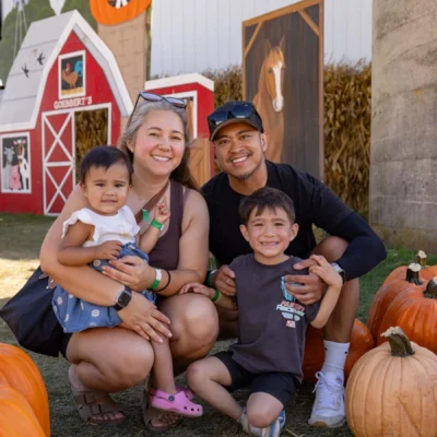A family of four poses among pumpkins outside a barn decorated with fall and Halloween-themed props during daytime.