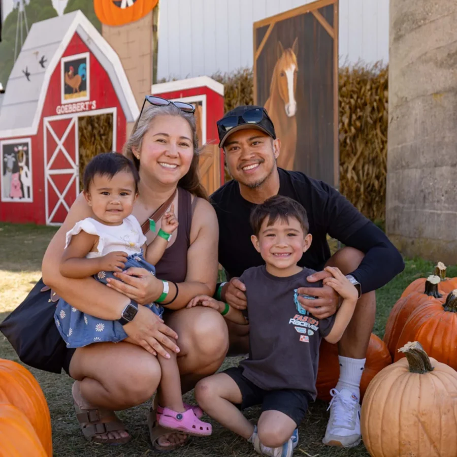 A family of four poses among pumpkins outside a barn decorated with fall and Halloween-themed props during daytime.