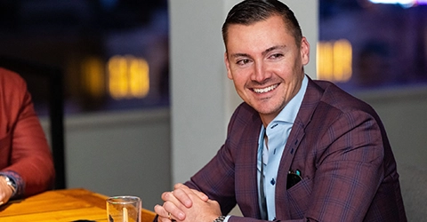 A man in a checked suit jacket sits at a table, smiling, with his hands clasped. There is a glass of water in front of him and blurred lights in the background.