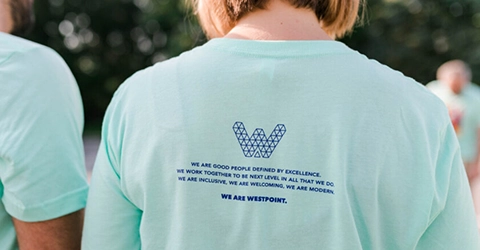 Person wearing a light blue T-shirt with the Westpoint logo and a printed statement about excellence, teamwork, inclusivity, and modernity on the back.