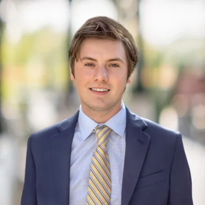 A young man in a blue suit and striped yellow tie stands outdoors, smiling at the camera with a blurred background.