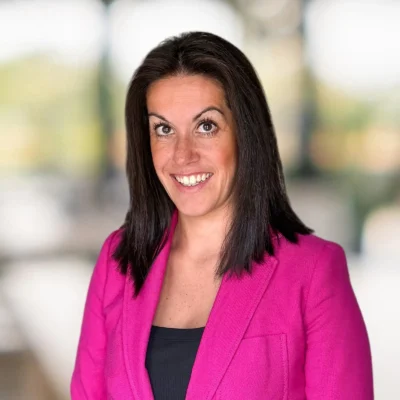 Woman with straight dark hair wearing a bright pink blazer and black top, smiling at the camera with a blurred background.