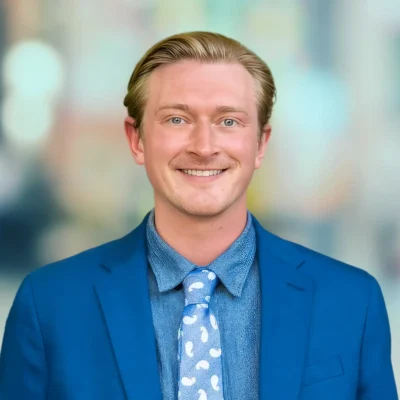 A man wearing a blue suit and blue patterned tie smiles at the camera against a blurred colorful background.