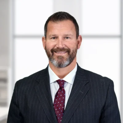 A man with short brown hair and a beard, wearing a dark pinstripe suit, white shirt, and maroon polka-dot tie, stands in a bright office setting.
