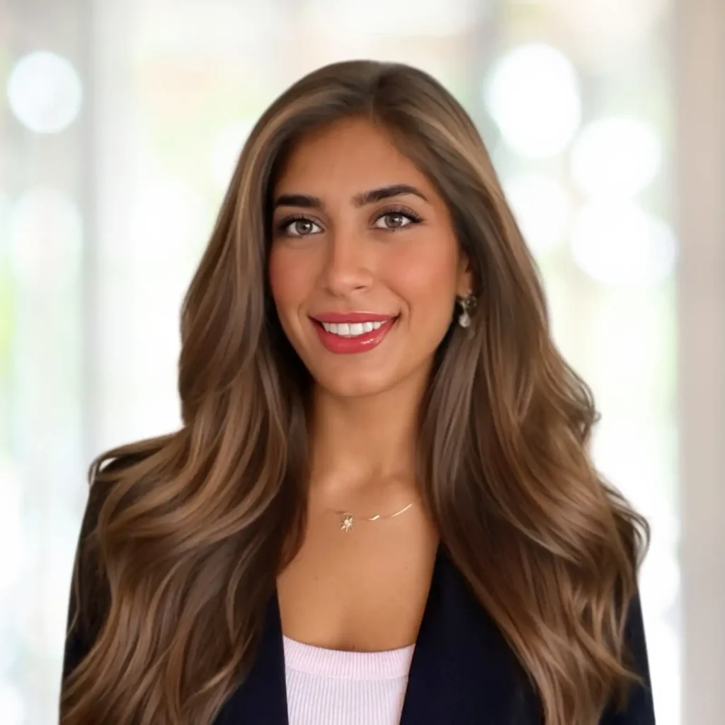 A woman with long, wavy brown hair, wearing a blazer over a light top, smiles at the camera against a blurred light background.