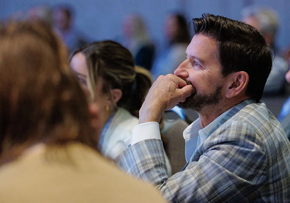 A man in a plaid suit jacket sits among a group, resting his chin on his hand, appearing attentive during an indoor event or meeting.
