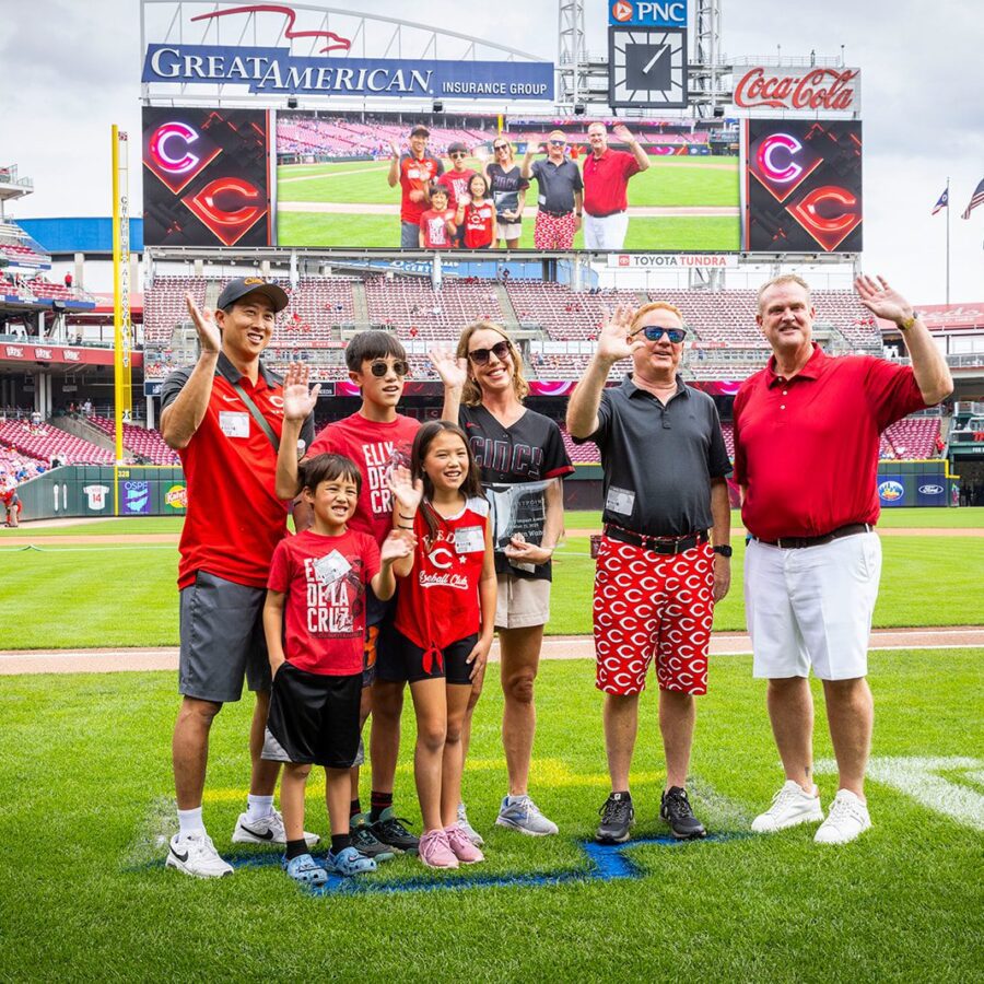 A group of seven people pose and wave on a baseball field, with a stadium and a large screen in the background displaying their image.