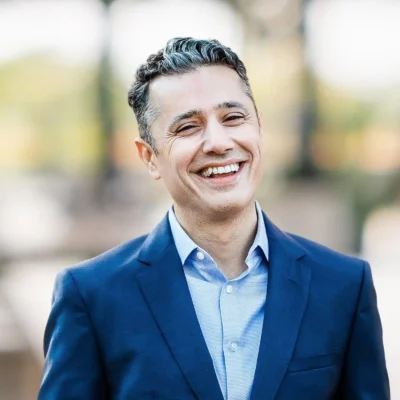 A man in a blue suit and light blue shirt is smiling outdoors with a blurred background.