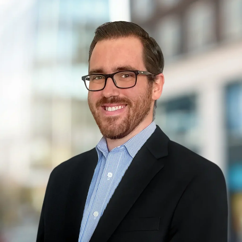 A man with short brown hair, glasses, and a beard is wearing a dark blazer and light blue collared shirt, smiling in front of a blurred urban background.