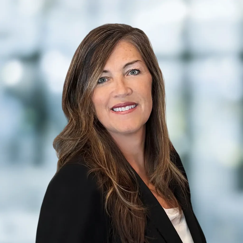Woman with long brown hair wearing a black blazer and white top, smiling in front of a blurred, light blue background.