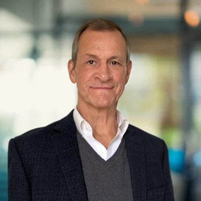 A middle-aged man wearing a dark blazer, grey sweater, and white shirt stands indoors with a blurred office background.