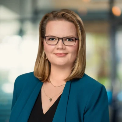 A woman with shoulder-length blonde hair, glasses, and a teal blazer poses for a professional headshot in an office setting.