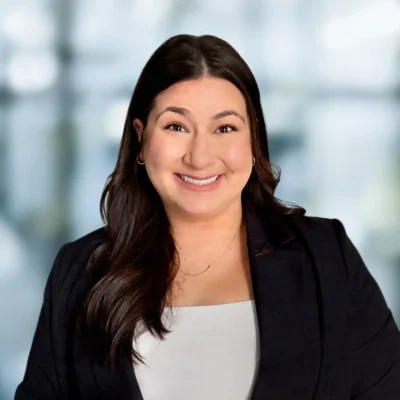 A woman with long dark hair, wearing a black blazer and white top, smiles at the camera with a blurred office background.