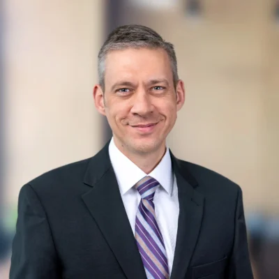 A man in a dark suit, white shirt, and striped tie stands in front of a blurred, neutral background, smiling slightly.