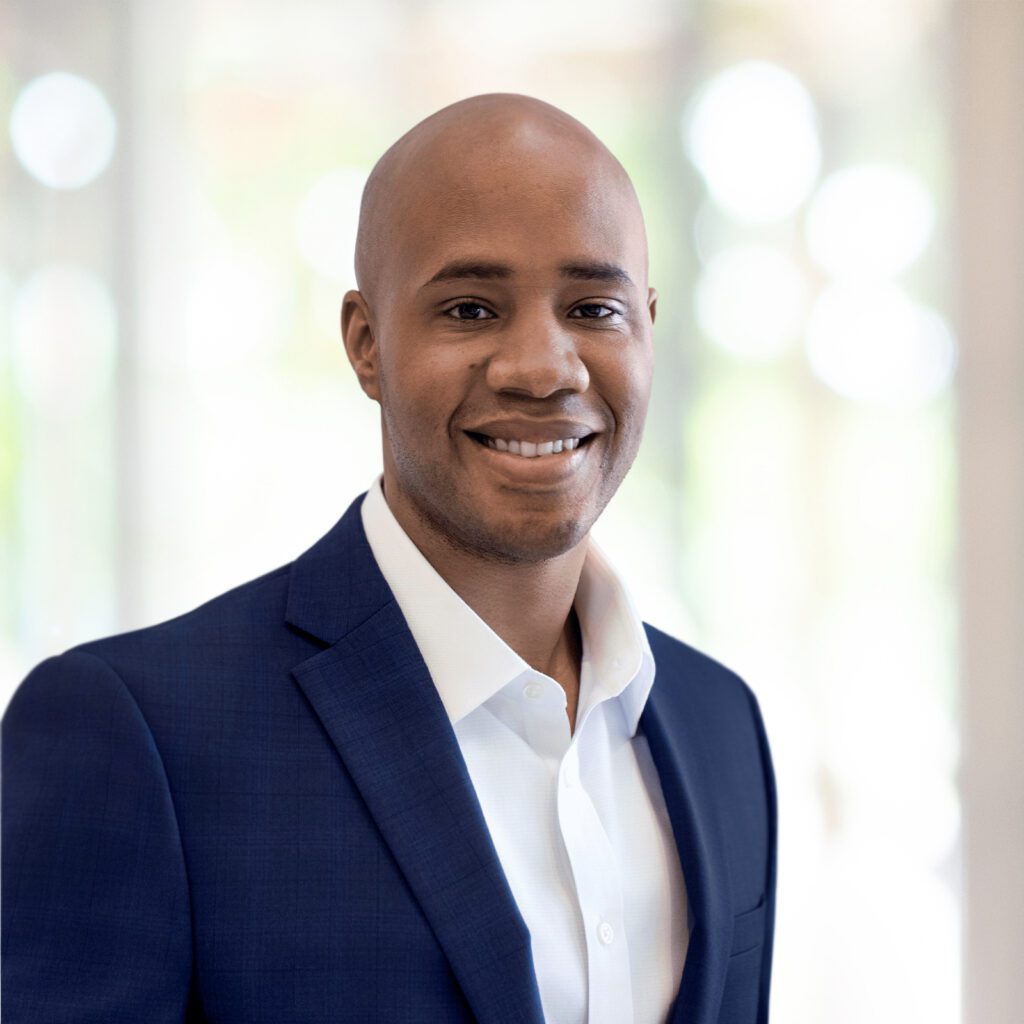 A man in a blue suit jacket and white shirt smiles at the camera, standing in a brightly lit indoor setting with a blurred background.