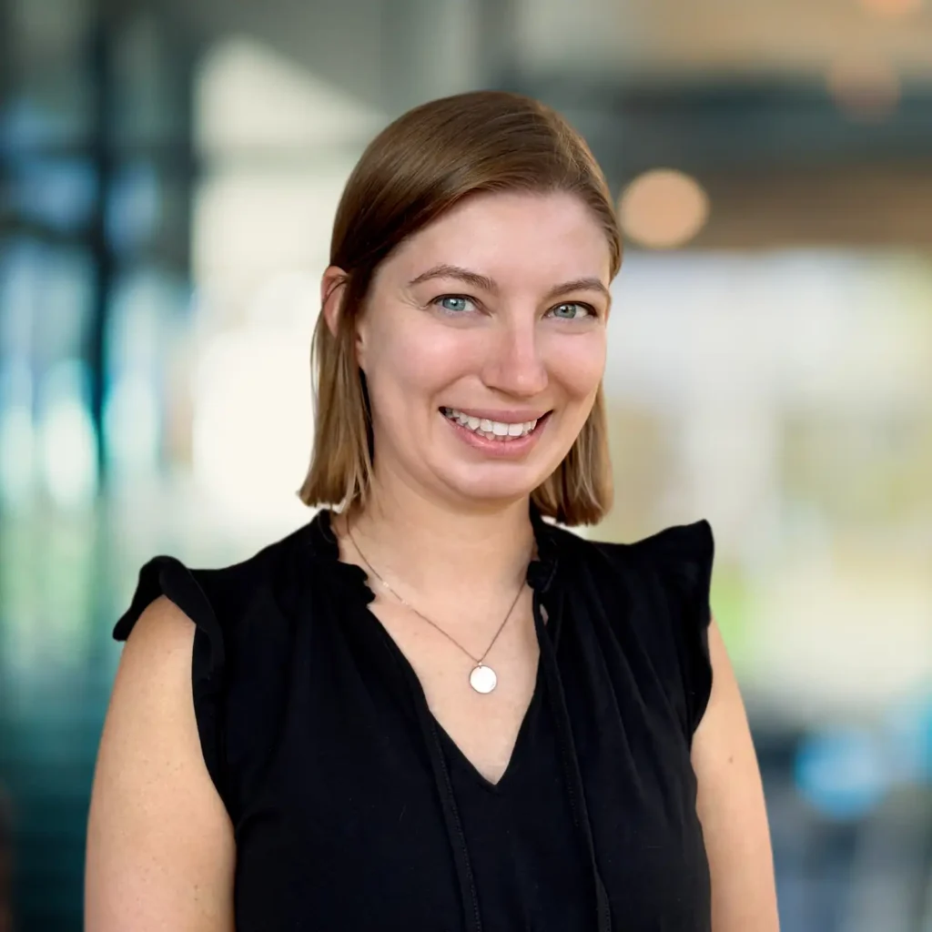 A woman with short light brown hair, wearing a black sleeveless top and a necklace, smiles at the camera in a brightly lit indoor setting.